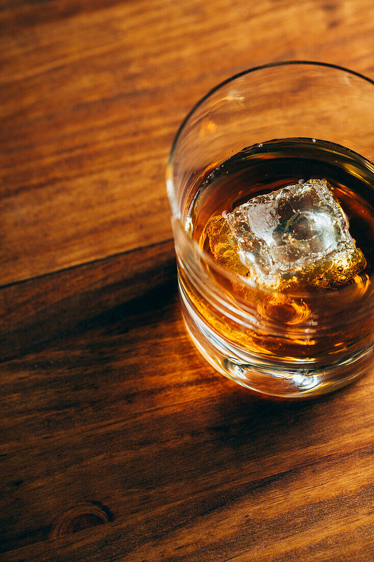 From above glass cup with cold whiskey and cube of ice placed on lumber table in dark room