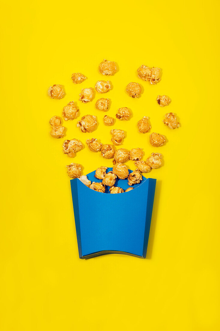 Top view of bright blue paper bag with delicious scattered sweet caramel popcorn on yellow background