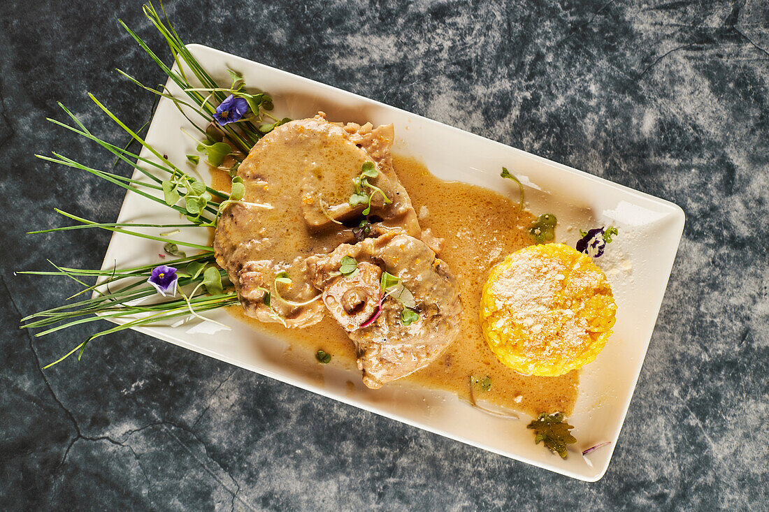 Von oben köstliche Ossobuco Gericht mit Gemüse und Reis-Sauce garniert mit Blumen auf rechteckigen Teller auf Marmortisch während des Mittagessens im Restaurant