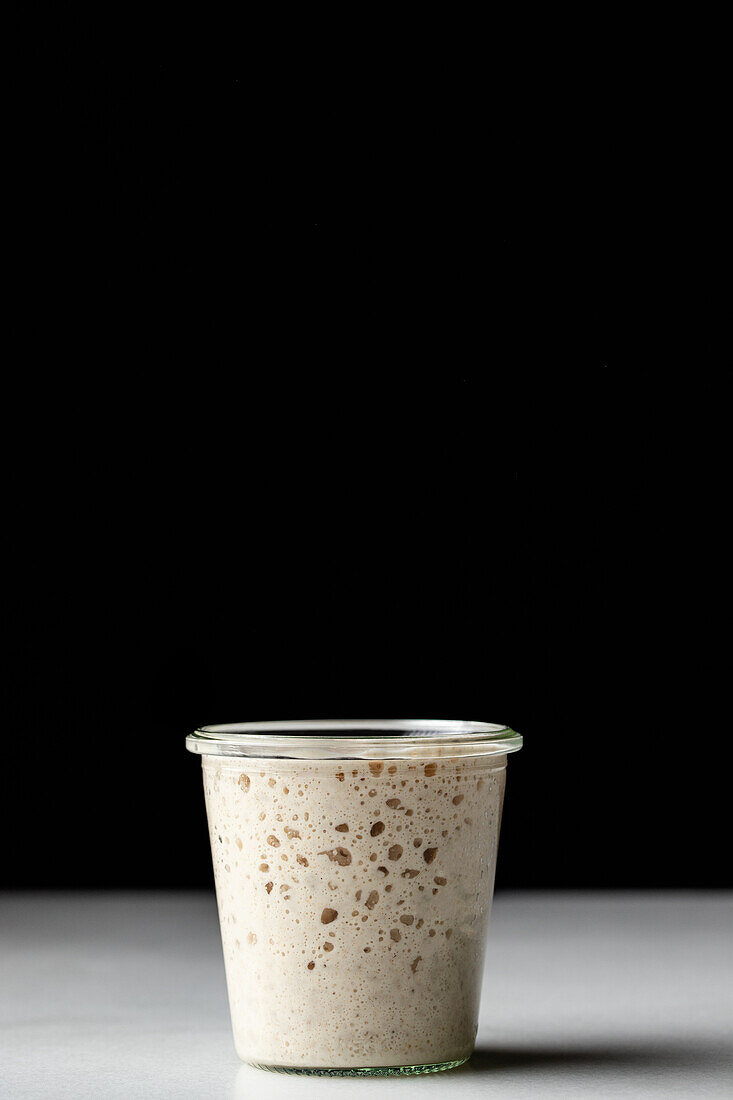 Glass white cup with sourdough starter covered with glass transparent lit placed on surface against black background