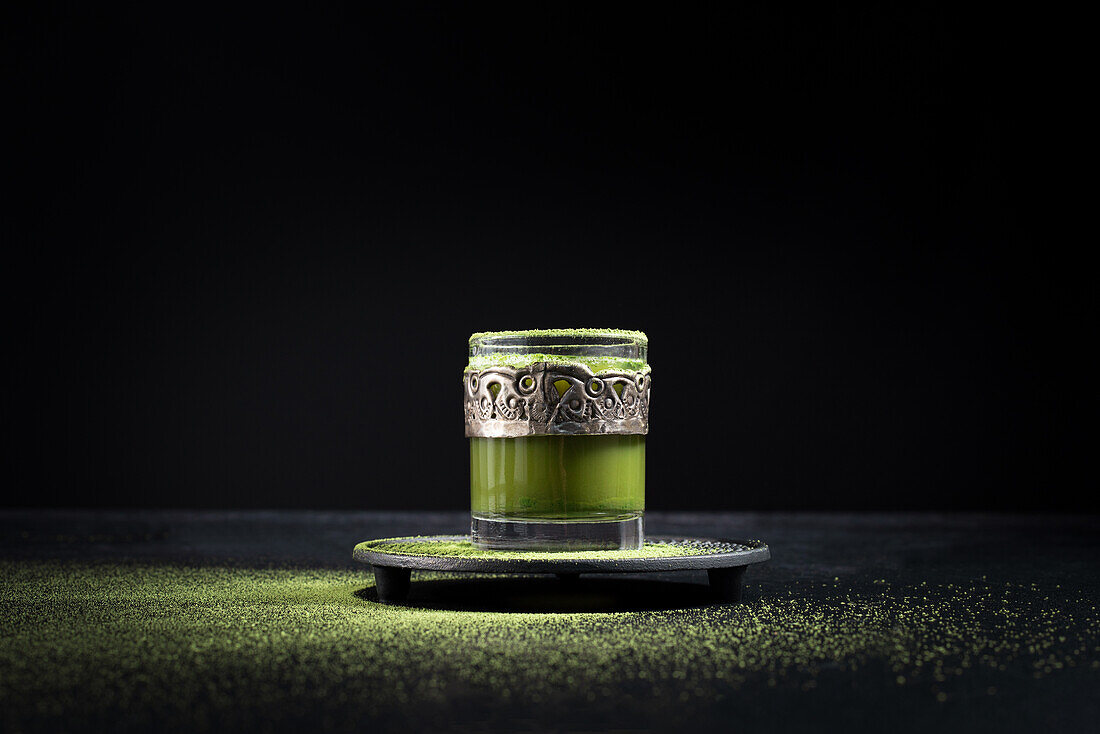 Gesunder grüner Matcha-Kräutertee, serviert in einer Glastasse mit Metallverzierung auf einer Untertasse, bestreut mit Pulver auf einem schwarzen Tisch