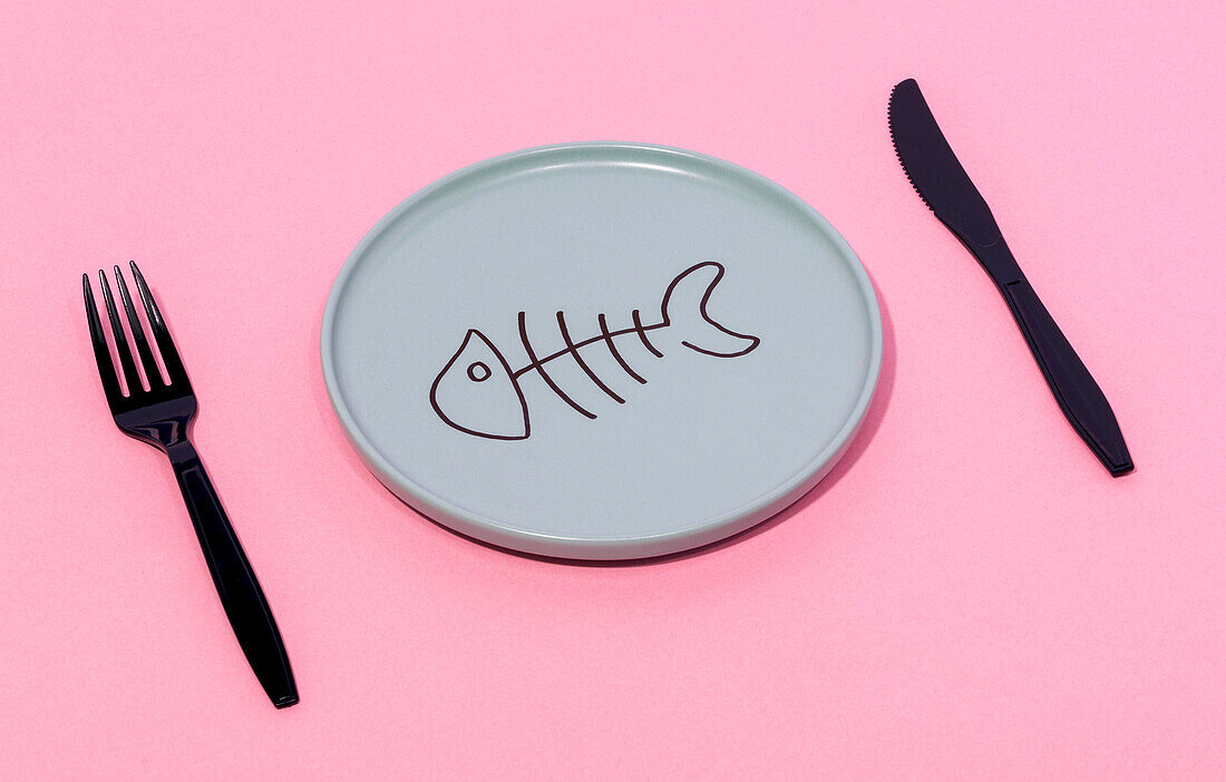 From above of gray ceramic plate with painted fish skeleton served on pink surface with black fork and knife