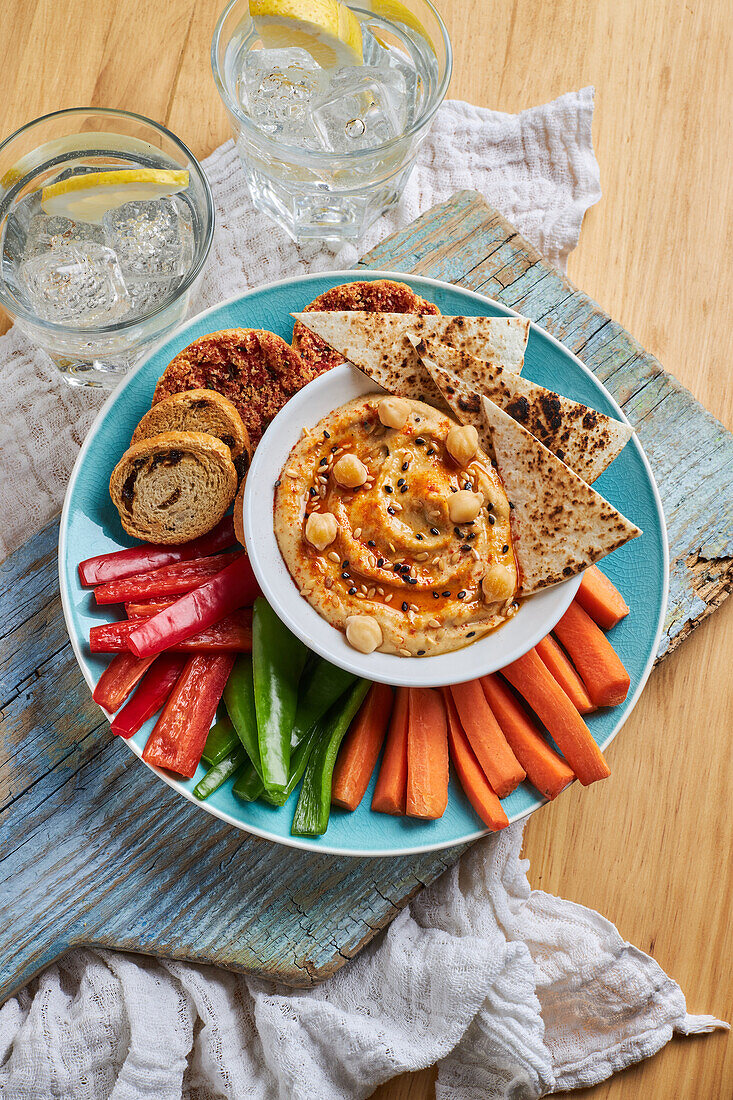 Top view of yummy hummus with tortilla chips served on plate with croutons and assorted vegetables on rustic cutting board near glasses of cold lemonade on wooden table with napkin