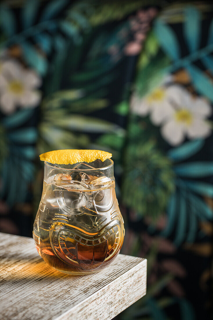 Tiki glass mug with booze placed on edge of wooden table in room with colorful curtain on blurred background