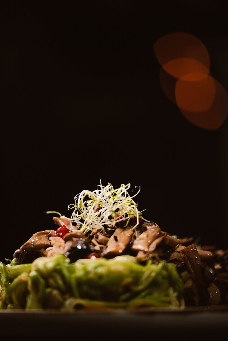 Low angle of yummy vegan dish with zucchini spaghetti and sauteed mushroom slices covered with red berries and alfalfa sprouts on dark background