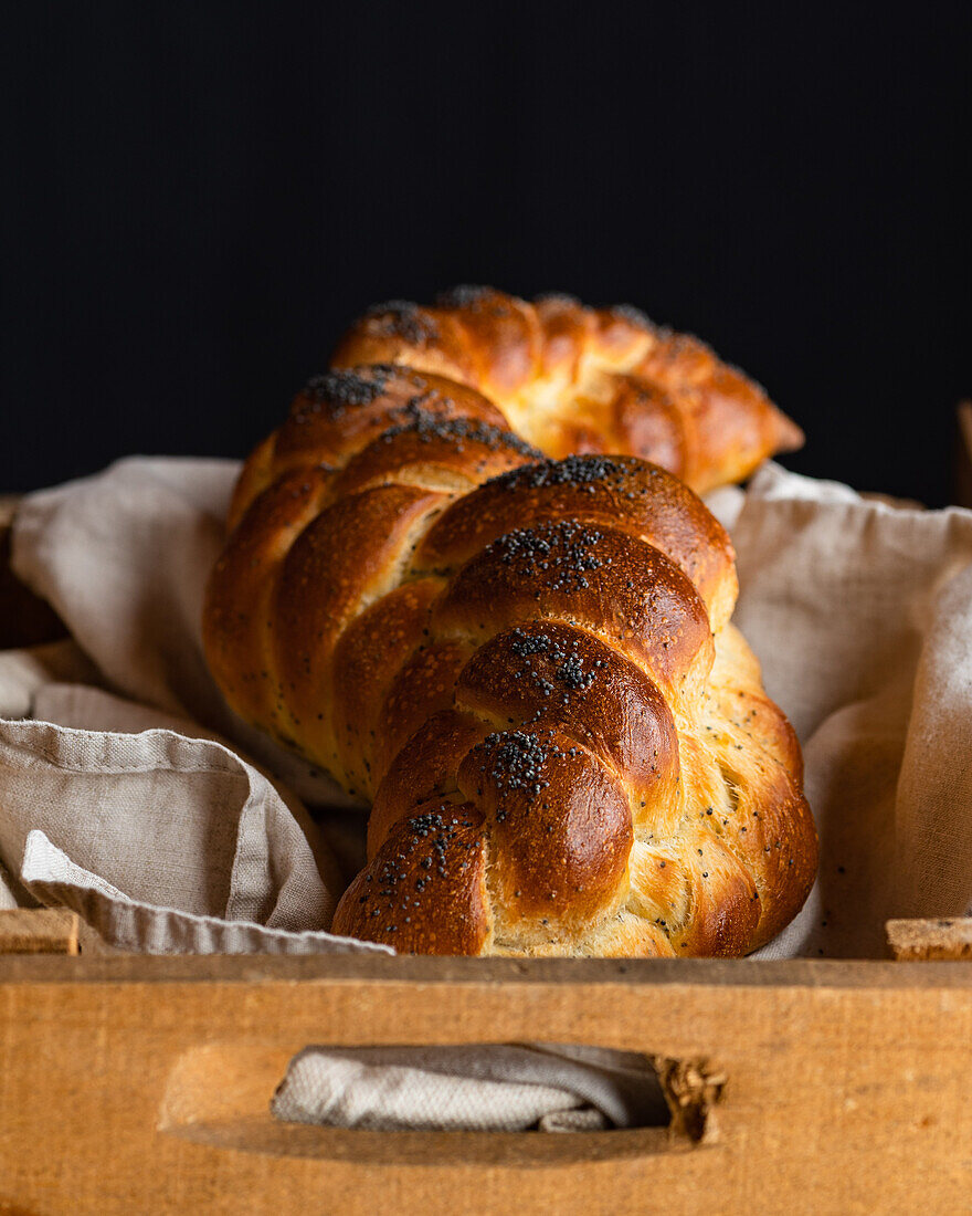 Served golden brown bread braids challah with poppy seeds on white napkin in wooden box