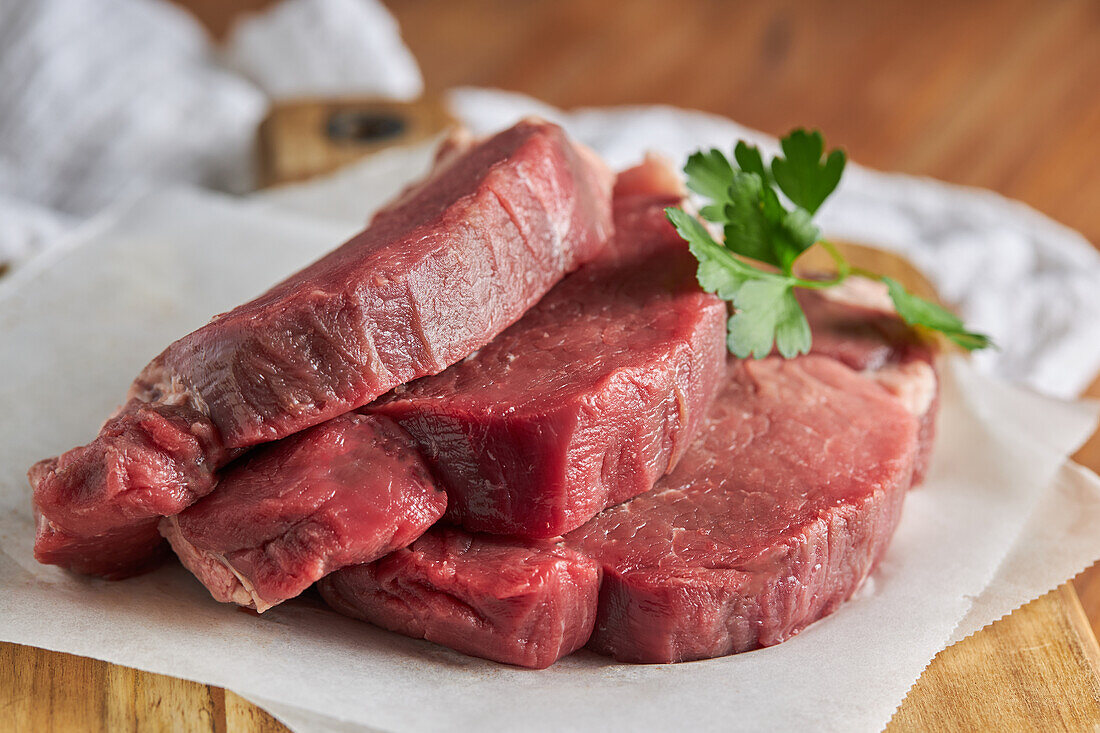 From above slices of fresh uncooked beef fillet with green parsley placed on wooden chopping board with parchment paper in kitchen