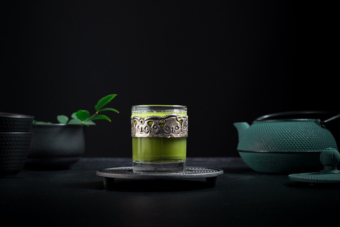 Still life composition with traditional oriental matcha tea served in glass cup with metal ornamental decor on table with ceramic bowls and fresh green leaves against black background