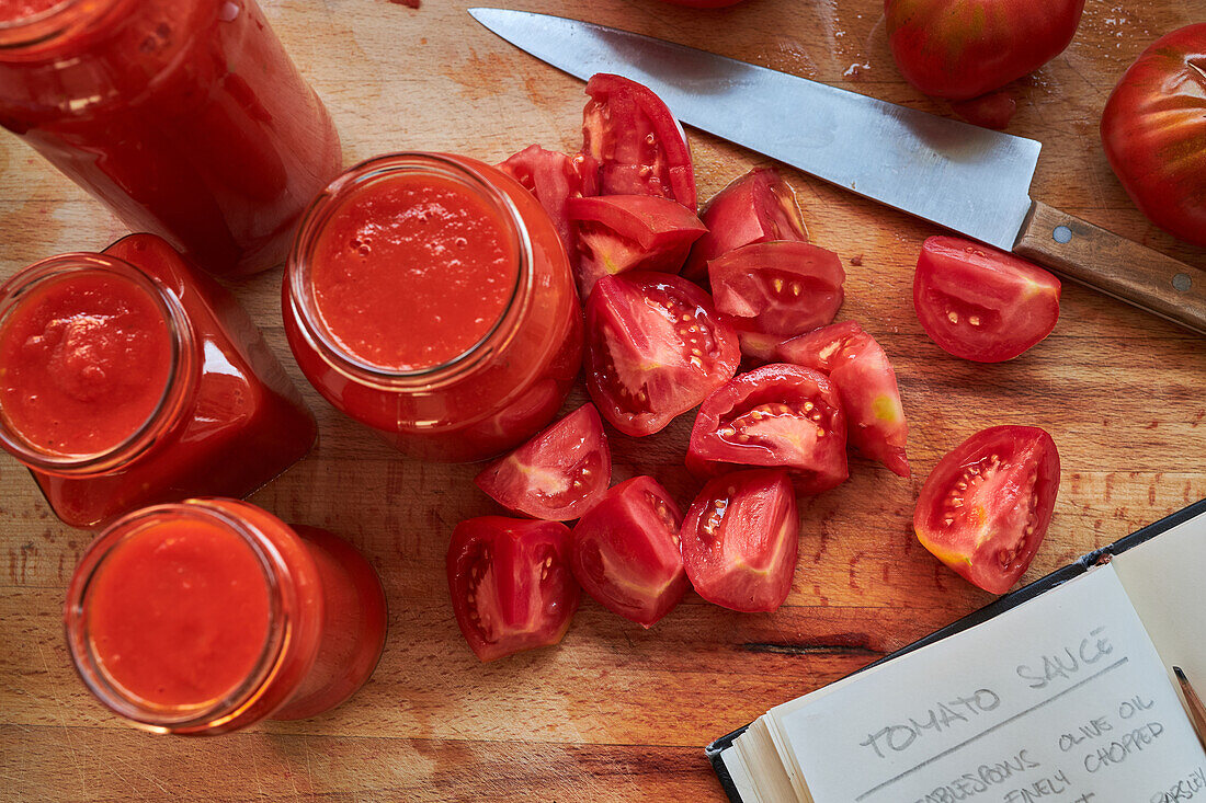 Reife rote Tomaten und Gläser mit Soße stehen auf dem Tisch neben einem Notizbuch mit Rezept in der Küche