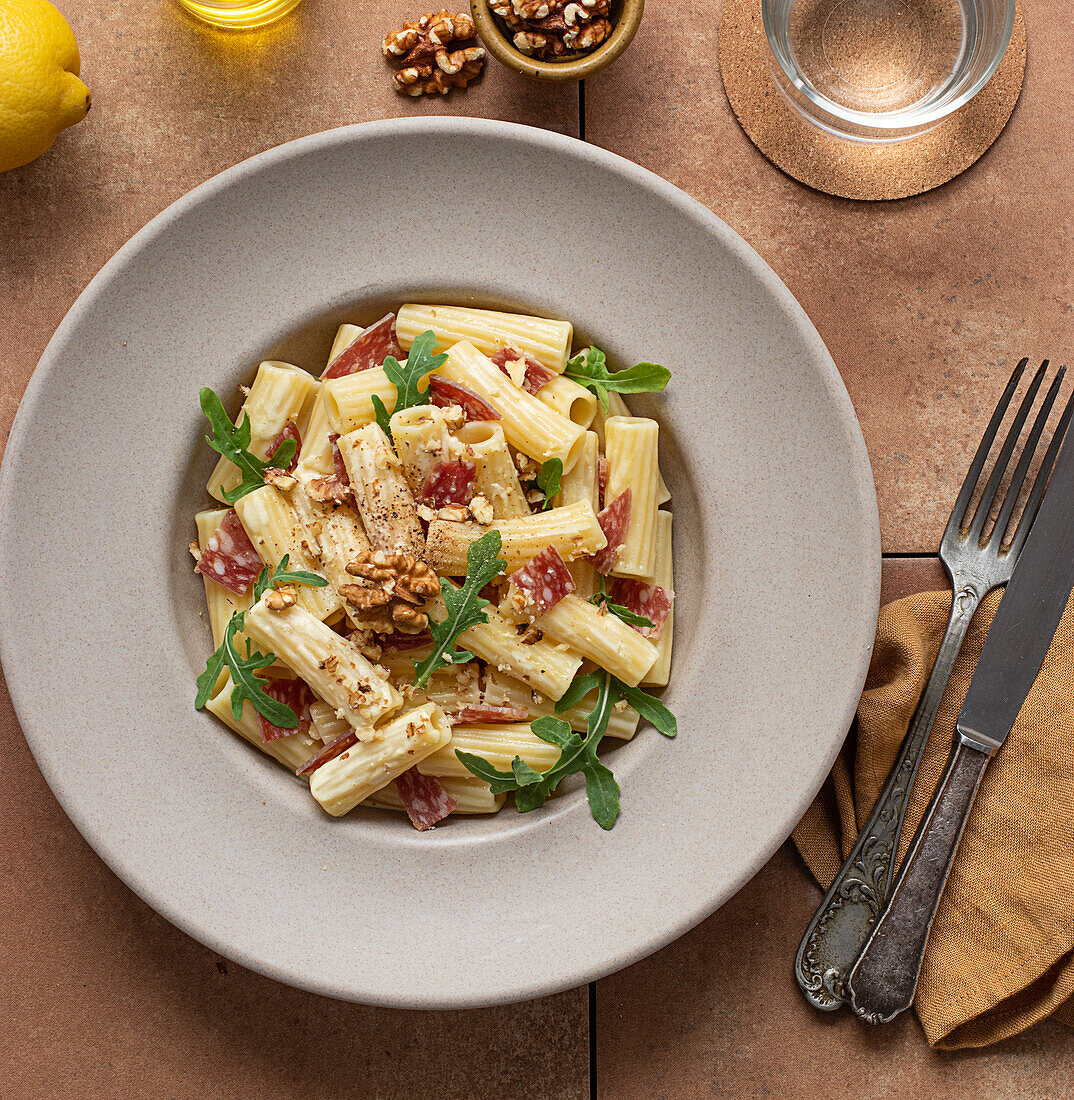 Nudelgericht mit Rucola, Salami und Walnüssen von oben auf einem Tisch, umgeben von Öl, Zitrone und Besteck