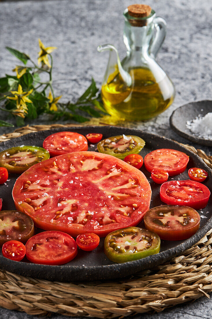 Delicious sliced tomatoes in cast iron plate placed on wicker napkin near plate with sea salt and jug of olive oil on concrete table