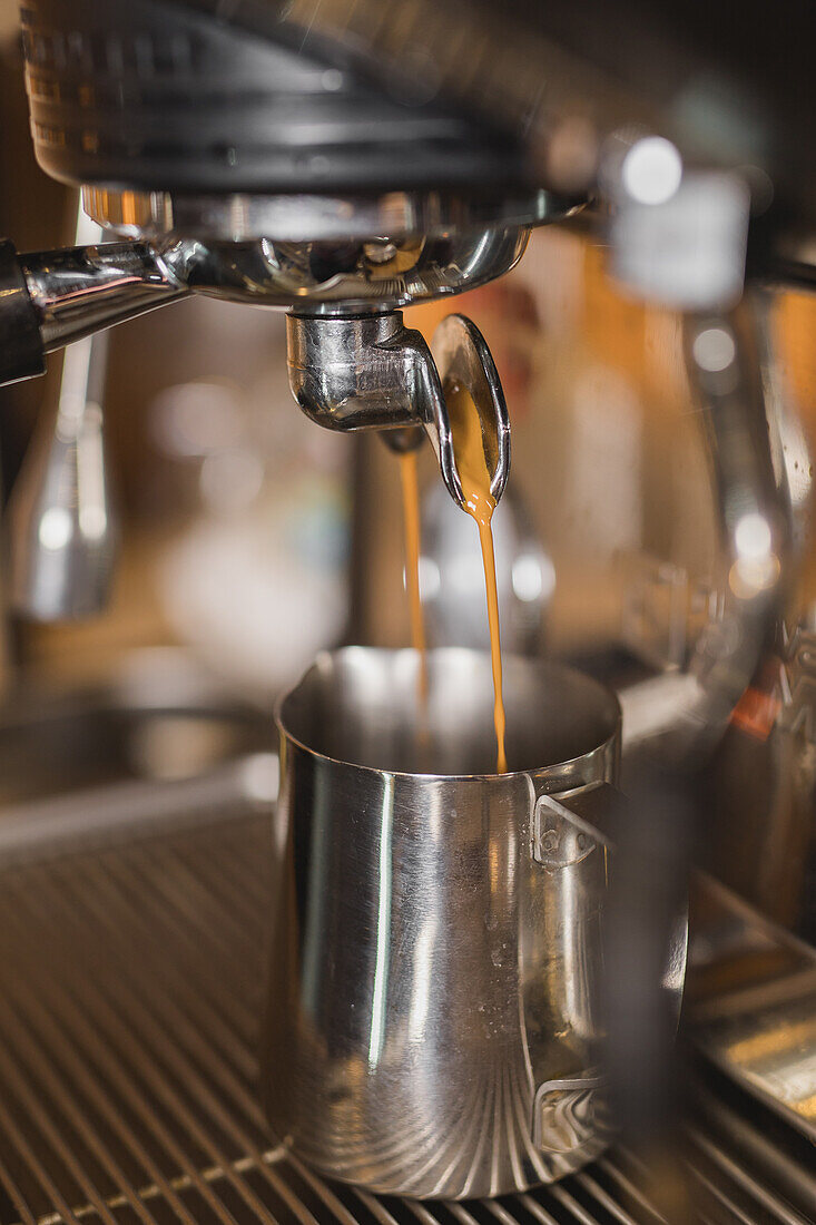 Freshly brewed espresso pouring from portafiller in professional coffee machine into stainless milk pitcher