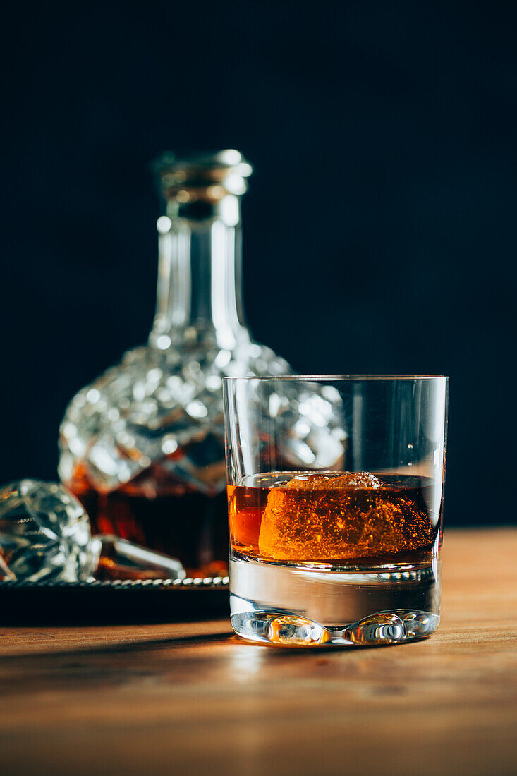 Glass of cold whiskey with ice placed on wooden table near decanter on black background