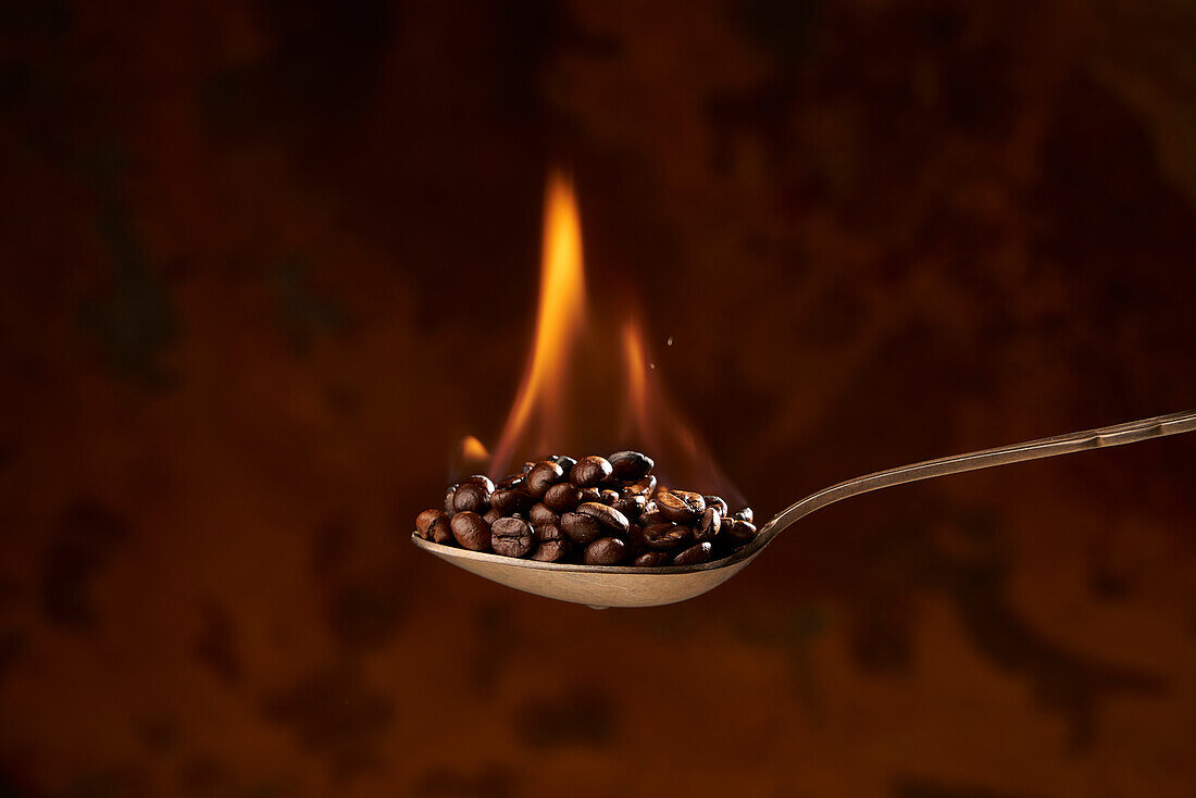 Nahaufnahme eines Löffels voll mit einem Haufen gerösteter Kaffeebohnen mit Feuer auf braunem Hintergrund