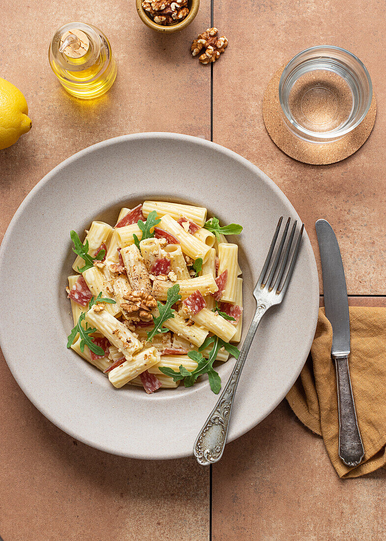 From above dish of pasta with arugula, salami and walnuts on a table surrounded by oil, lemon and cutlery