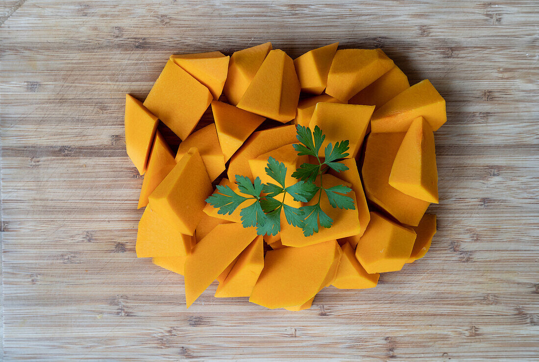 Top view of pile of pieces of ripe pumpkin garnished with fresh sprig of parsley placed on wooden table