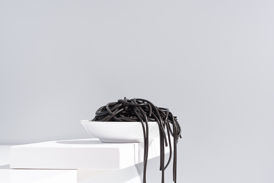 Minimalist studio with black squid ink spaghetti falling out from full ceramic bowl on white table