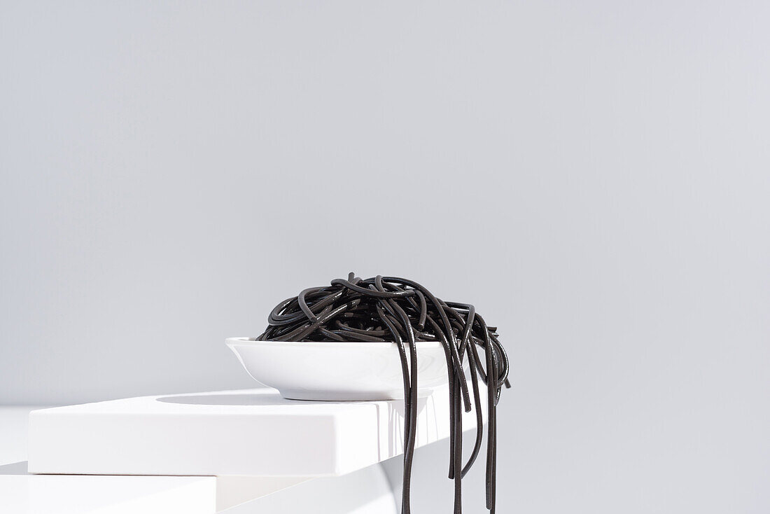 Minimalistisches Atelier mit schwarzen Tintenfischspaghetti, die aus einer vollen Keramikschüssel auf einen weißen Tisch fallen