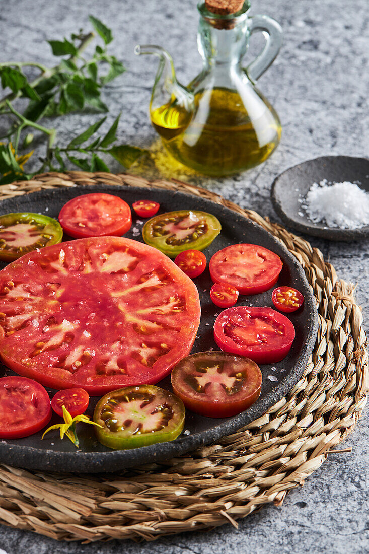 Delicious sliced tomatoes in cast iron plate placed on wicker napkin near plate with sea salt and jug of olive oil on concrete table