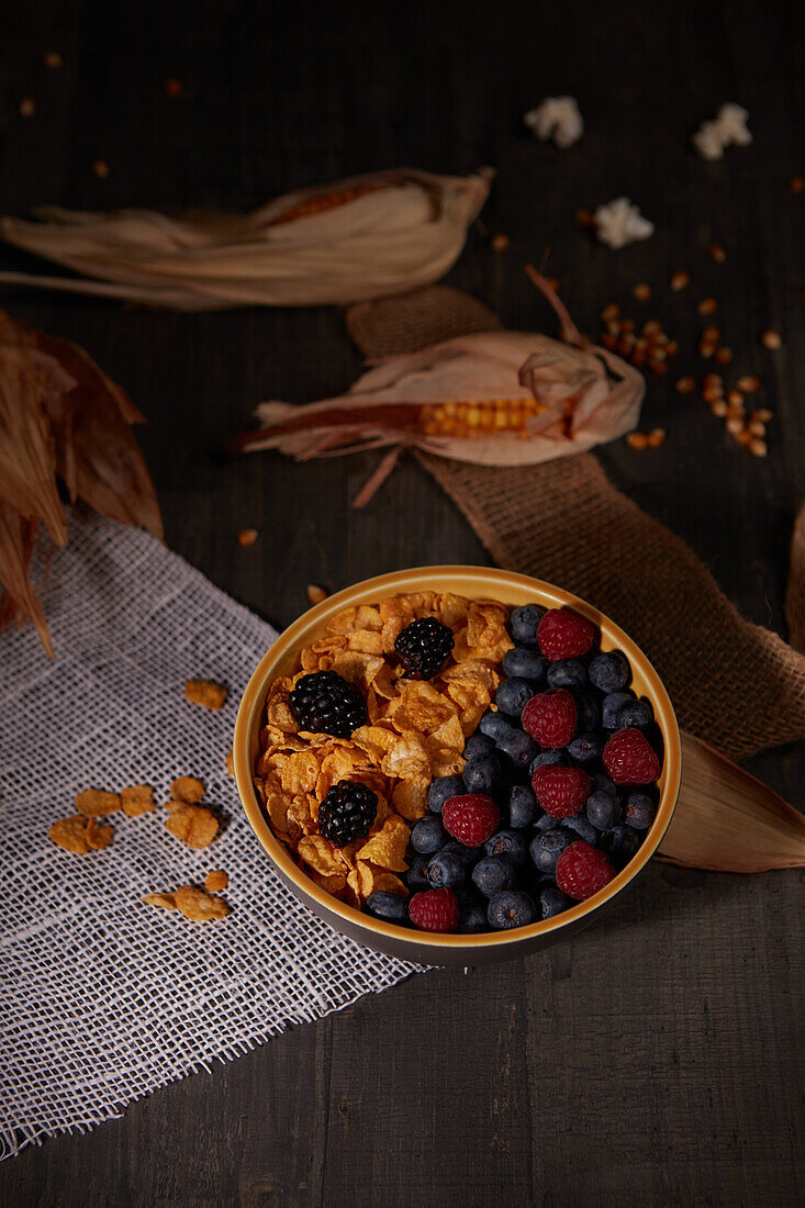 Von oben leckere Cornflakes mit frischen Waldbeeren in einer Schüssel neben Sackleinen beim Frühstück in einem dunklen Raum