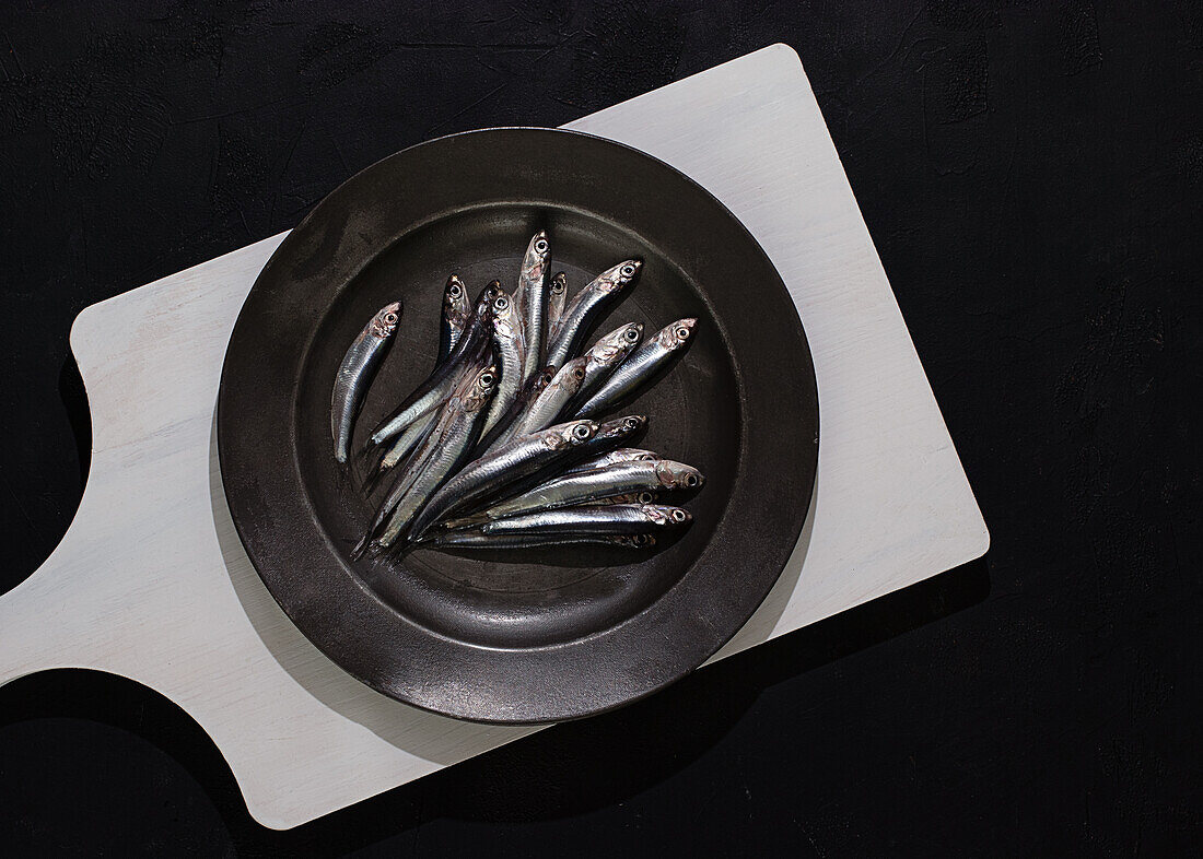 Top view of raw small fishes in plate on white board on black background