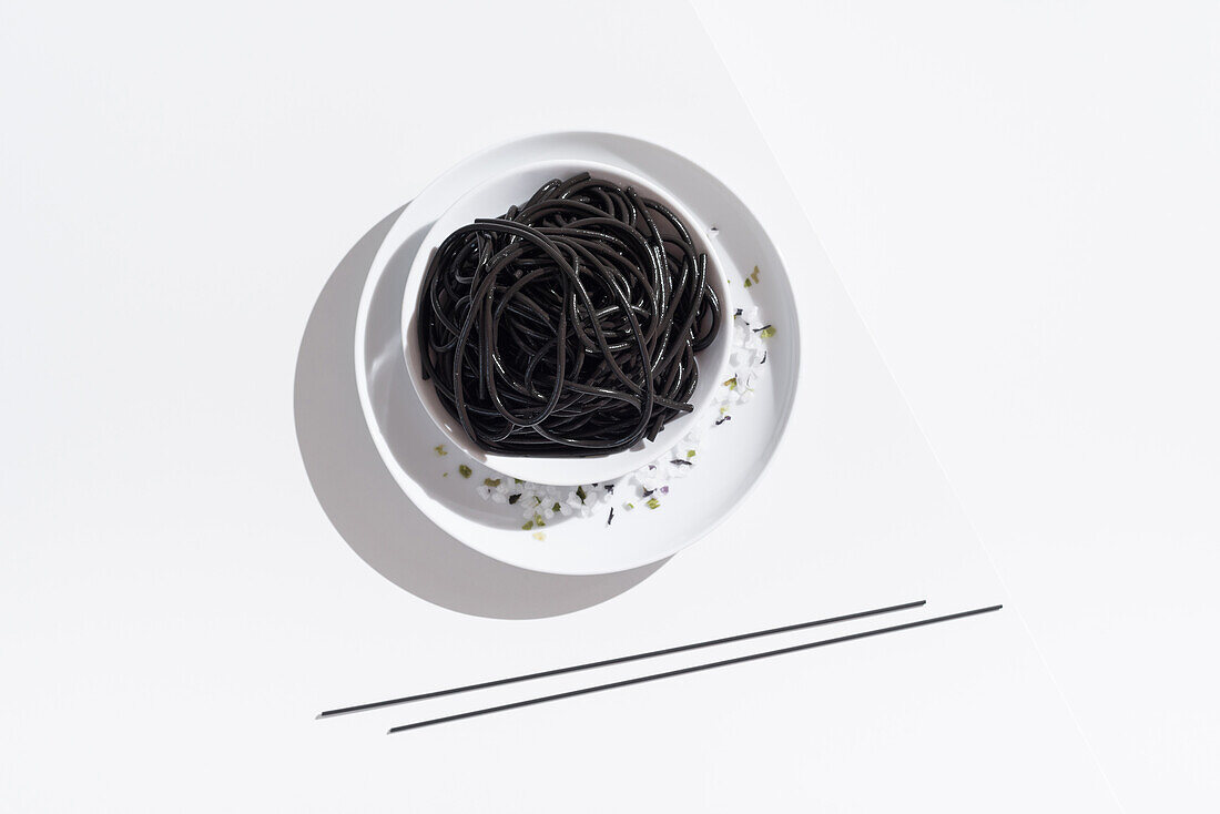 From above of ceramic bowl with delicious spaghetti with black squid ink with chopsticks on white background