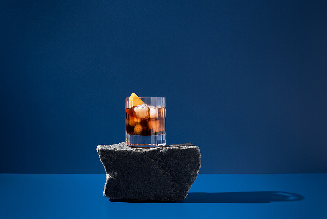 Glass of alcoholic cocktail made with red vermouth served with orange slices on stone in light studio with blue wall