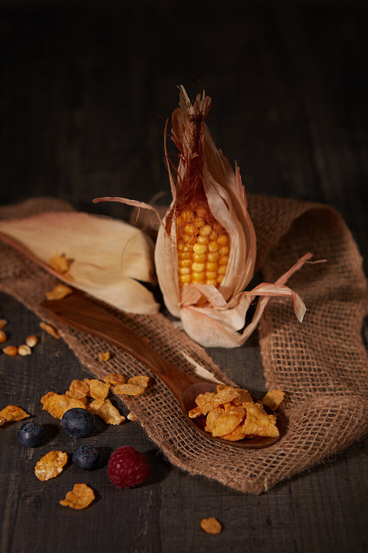 Frischer Mais und Cornflakes neben reifen Waldbeeren und einem Streifen Sackleinen auf einem Holztisch in einem dunklen Raum