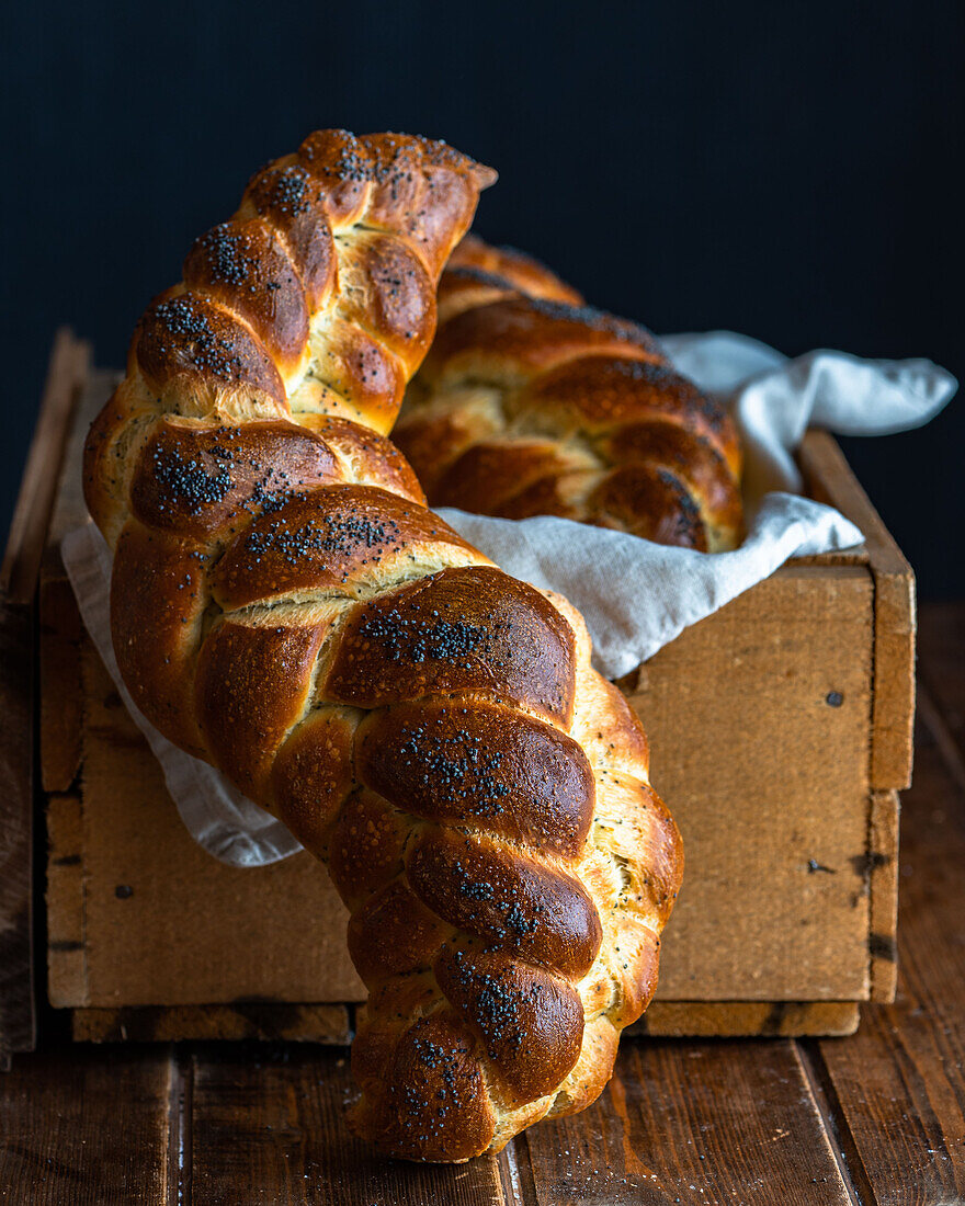 Served golden brown bread braids challah with poppy seeds on white napkin in wooden box