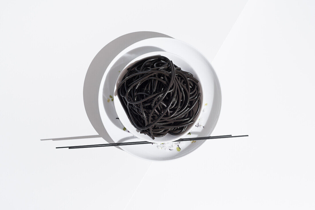 From above of ceramic bowl with delicious spaghetti with black squid ink with chopsticks on white background