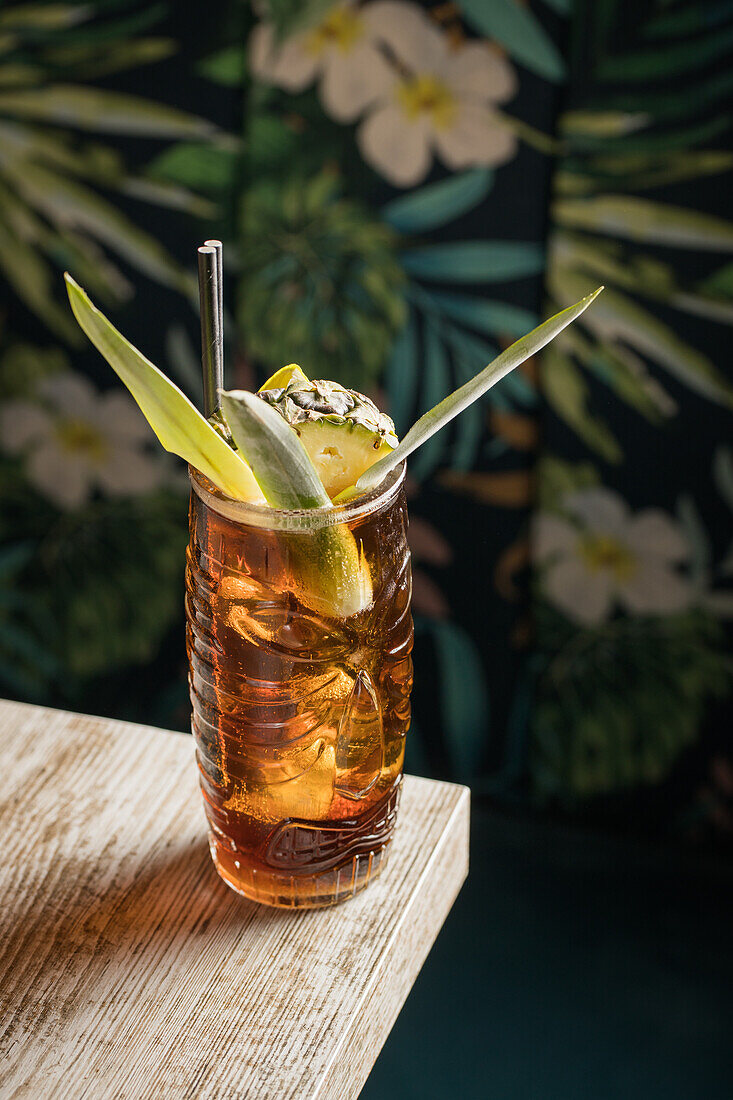 Tiki-Glasbecher von oben, gefüllt mit einem alkoholischen Getränk mit Strohhalm, dekoriert mit Früchten, auf der Kante eines Holztisches