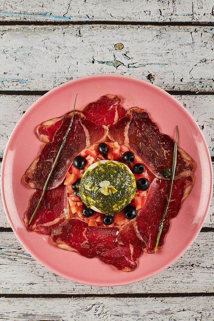 From above tomato salad with burrata and slices of prosciutto garnished with black olives and pesto sauce and served on plate on wooden table