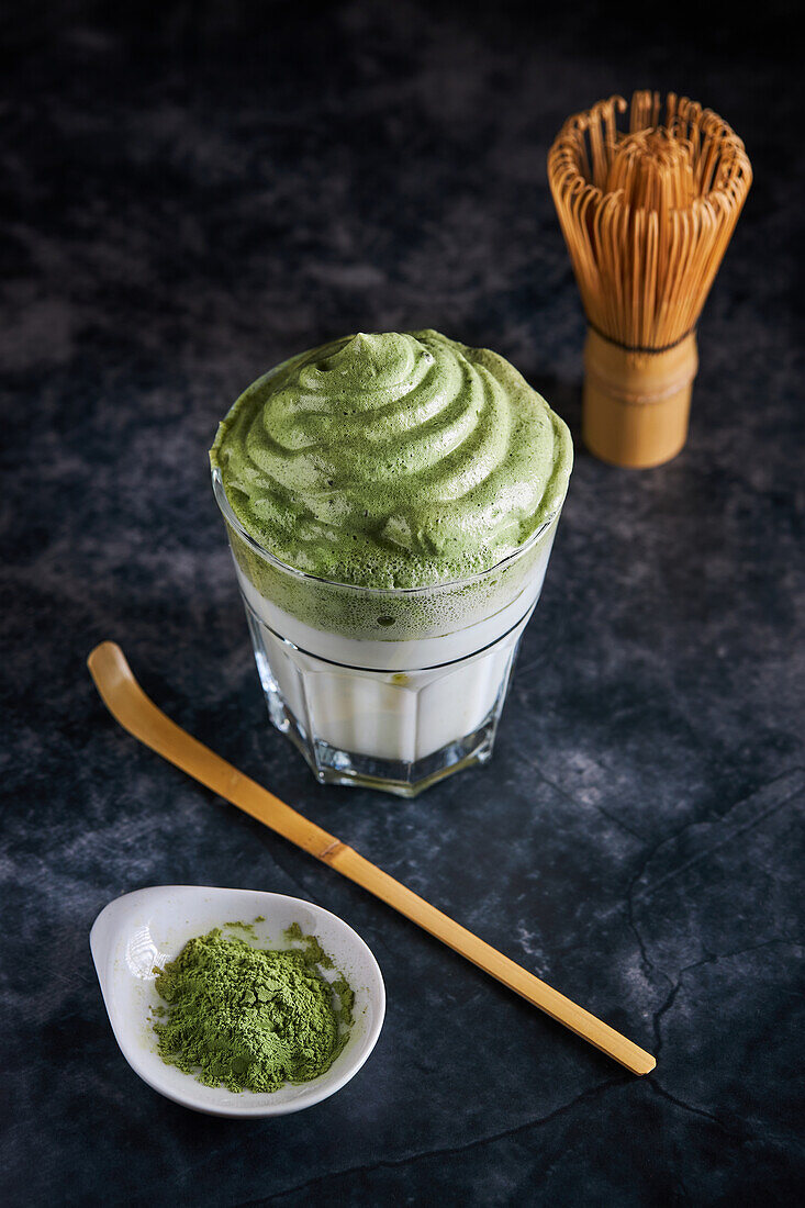 Blick von oben auf ein Glas mit frischem Dalgona-Kaffee mit Matcha und japanischen Teezeremonie-Accessoires, bestehend aus einem Bambusbesen namens Chasen und einer traditionellen Schöpfkelle namens Chashaku, die auf einer grauen Fläche platziert sind