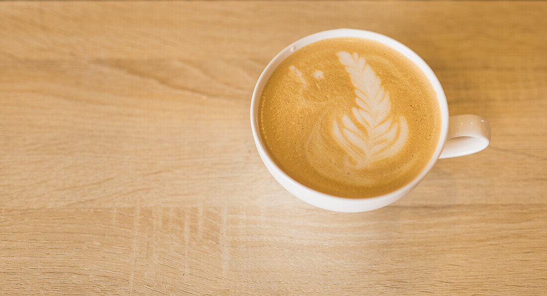 Blick von oben auf eine weiße Keramiktasse mit aromatischem, heißem Cappuccino, verziert mit Latte Art, auf einem Holztisch in einem Cafe
