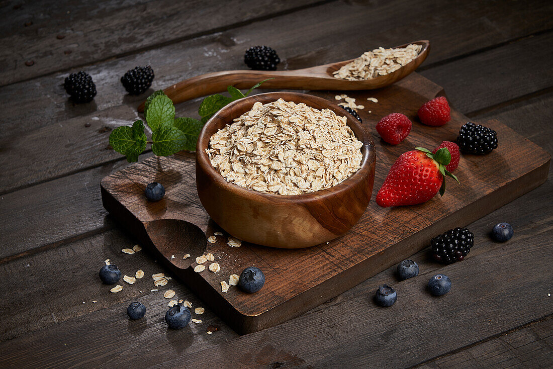 From above bowl with uncooked oatmeal served on wooden cutting board with various fresh berries and mint leaves in kitchen