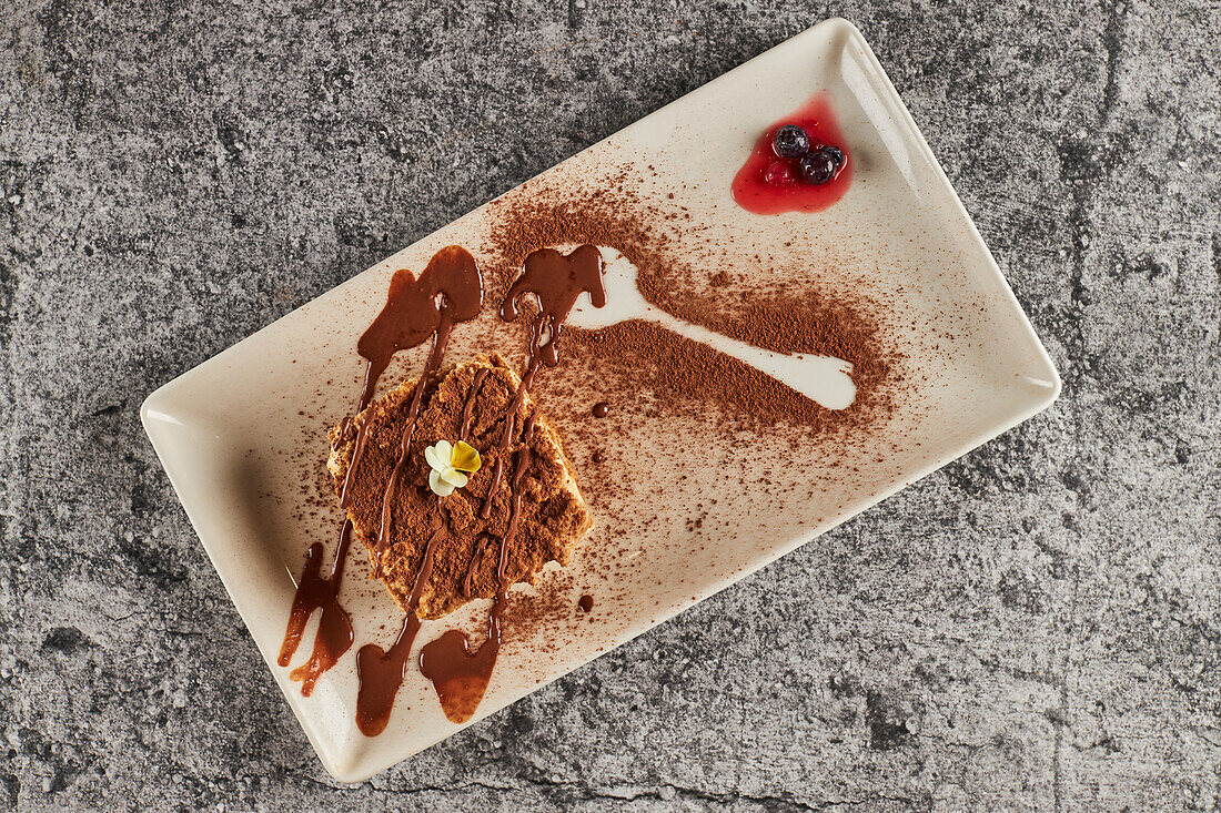 Top view of piece of delicious tiramisu dessert garnished with cocoa powder and chocolate sauce with flower and served on rectangular plate with berries on gray marble table in restaurant