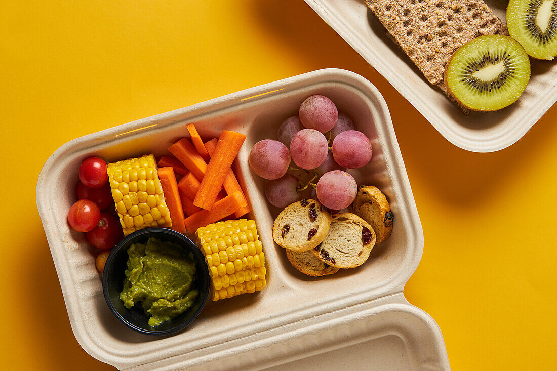 Von oben von Lunch-Boxen mit gesunden Lebensmitteln einschließlich Cracker Karotten-Sticks Trauben Kirschtomaten mit Kiwi Brokkoli Walnuss und Mandarine auf rosa Hintergrund