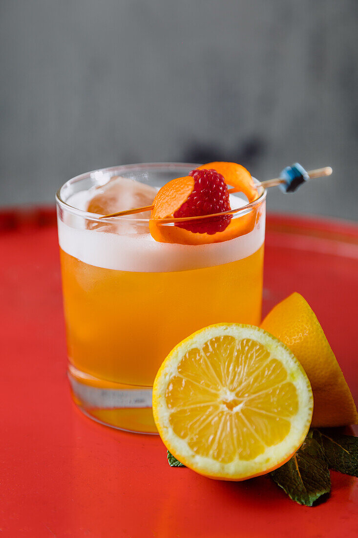 Kristallglas mit Amaretto Sour-Cocktail, garniert mit Orangenschale und Himbeeren, serviert mit einer halbierten Zitrone