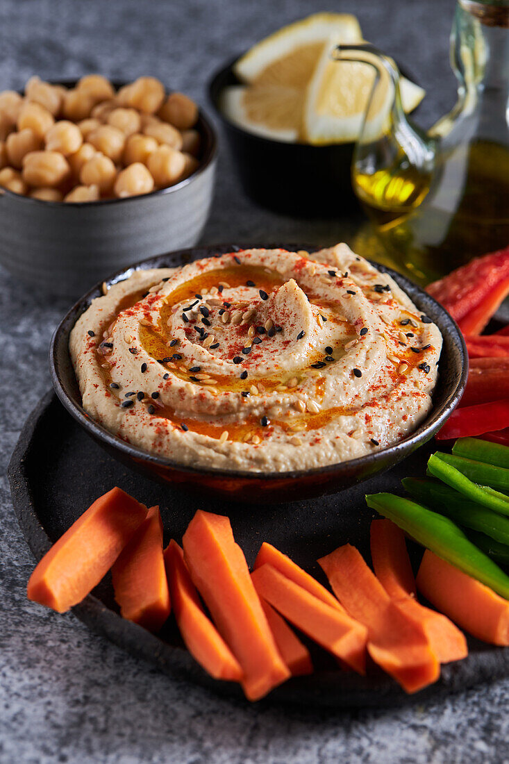 Von oben köstlicher Hummus und verschiedene Gemüsescheiben neben einer Flasche Öl und Schalen mit Kichererbsen- und Zitronenstücken auf einem grauen Marmortisch serviert