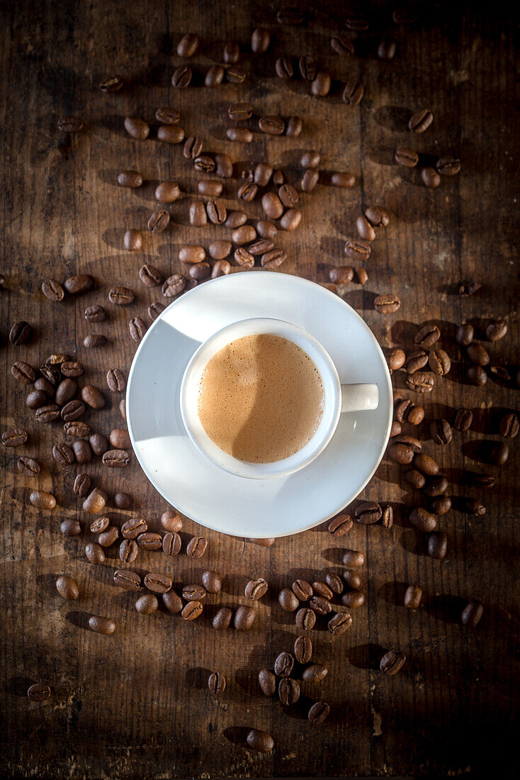 Top view of ceramic cup of coffee with foam among spilled roasted beans on wooden surface