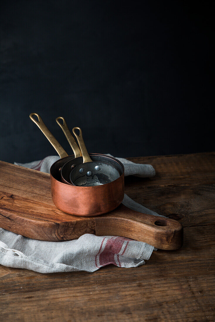Stack of copper saucepans placed on wooden cutting board and linen towel on rustic table on gray background