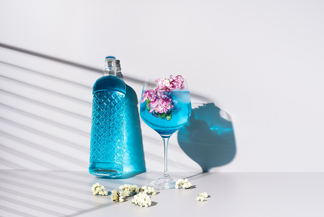 Glasflasche mit blauer Flüssigkeit in der Nähe eines Glases mit einem erfrischenden Cocktail mit Eis und Blumen auf einem Tisch vor weißem Hintergrund