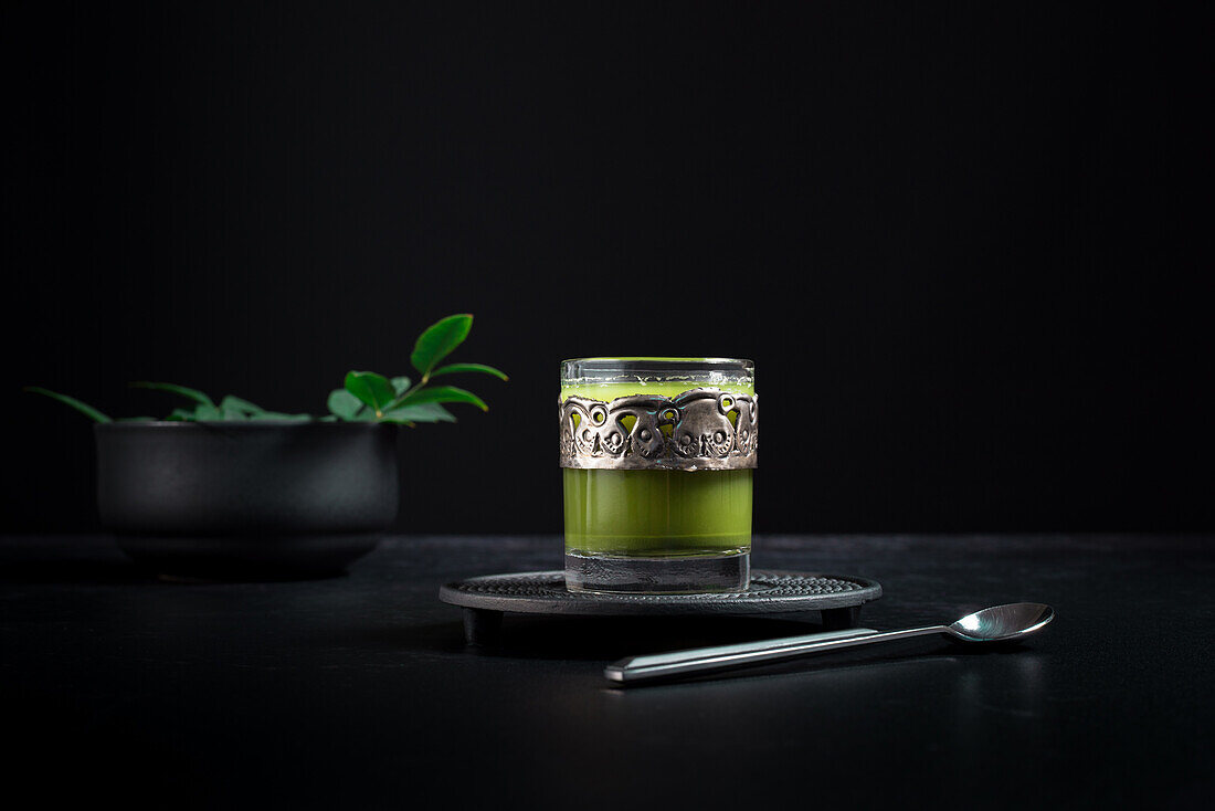 Still life composition with traditional oriental matcha tea served in glass cup with metal ornamental decor on table with ceramic bowls and fresh green leaves against black background