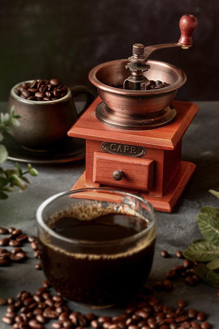 Glass of freshly brewed aromatic coffee on saucer placed on table with scattered beans against manual grinder