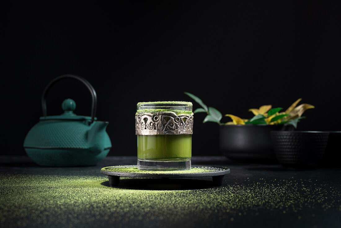 Stillleben Komposition mit traditionellem orientalischen Matcha-Tee in Glastasse mit Metallverzierung auf Tisch mit Keramikschalen und frischen grünen Blättern vor schwarzem Hintergrund