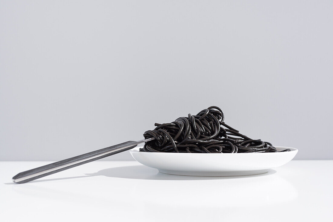 Stainless fork in full bowl of black spaghetti with cuttlefish ink on white table in studio on gray background