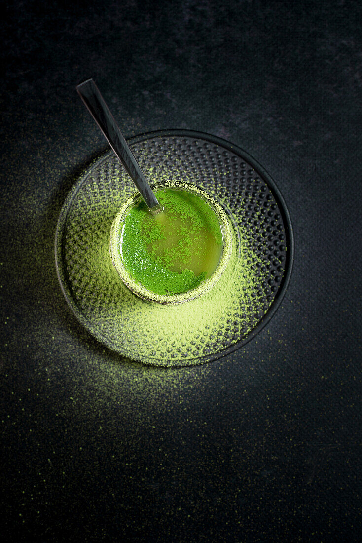 From above of healthy herbal green matcha tea served in glass cup with metal decoration on saucer sprinkled with powder on black table