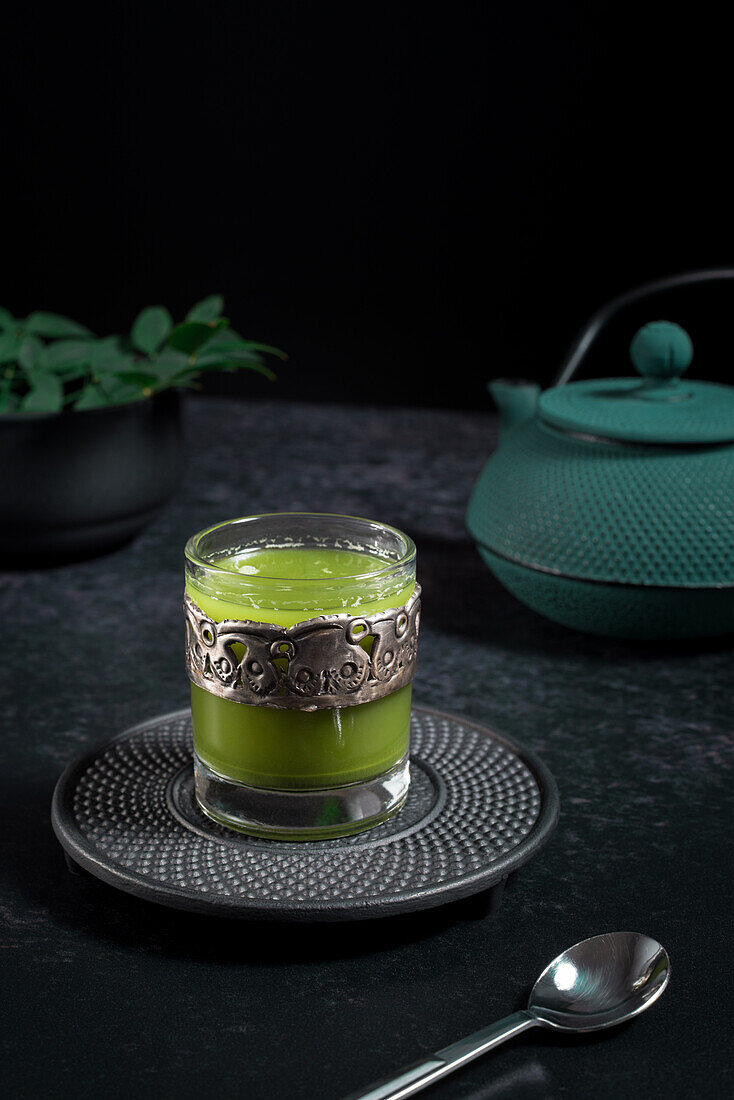 Still life composition with traditional oriental matcha tea served in glass cup with metal ornamental decor on table with ceramic bowls and fresh green leaves against black background