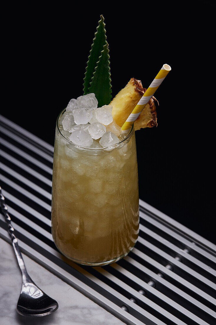 From above table with glass of yellow cocktail with ice cubes and refreshing cocktail garnished with spiky leaves and striped straw