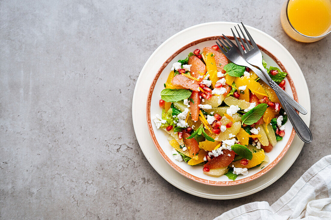 Draufsicht auf traditionellen griechischen Salat mit reifen Tomaten und Feta-Käse, gekrönt von Granatapfelkernen und Minzblättern auf grauem Hintergrund mit Orangensaft